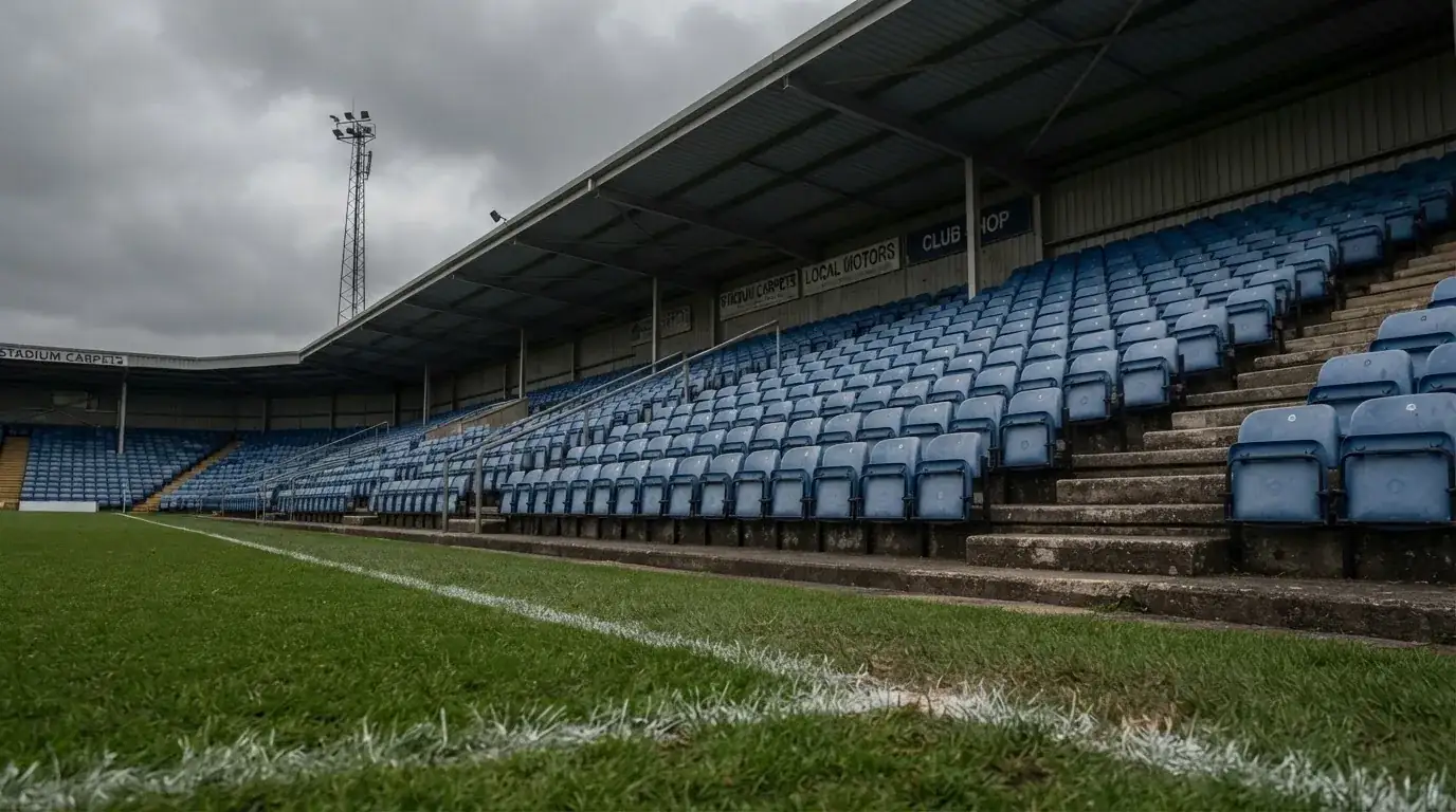 Rzędy pustych plastikowych krzesełek małego stadionu widziane z poziomu murawy pod pochmurnym niebem.