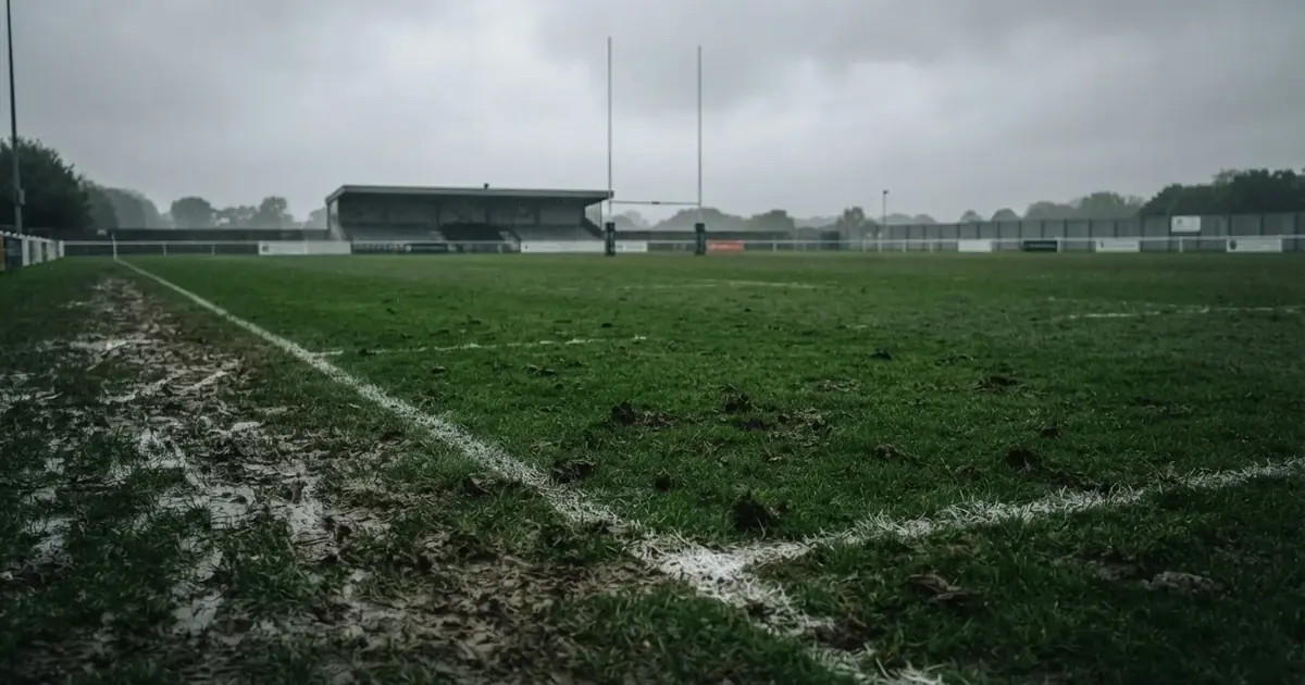 Boisko do rugby ze słupami bramki w oddali pod pochmurnym niebem, mokra trawa i ślady butów.