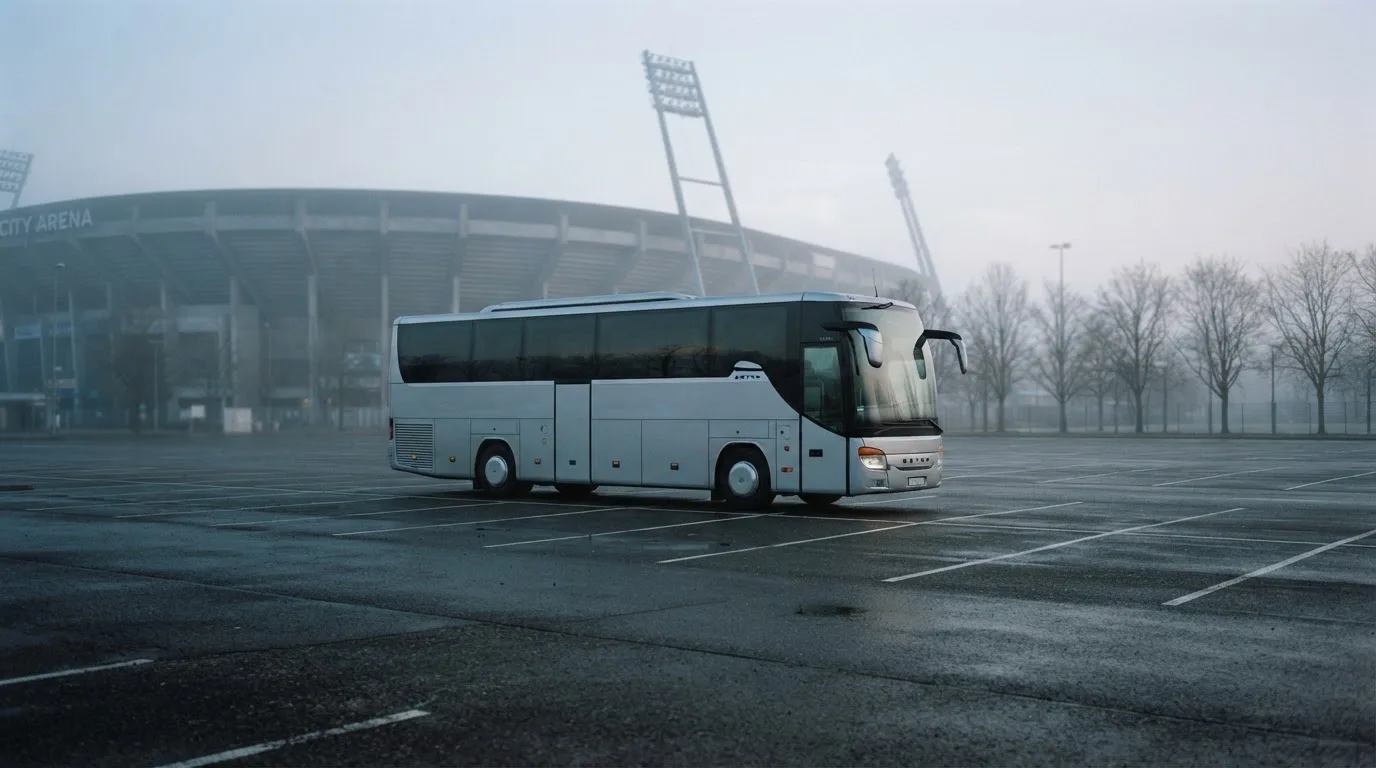 Autokar stoi na parkingu przed stadionem o świcie, rozmyte kontury trybun w tle, pusty plac i mokry asfalt.